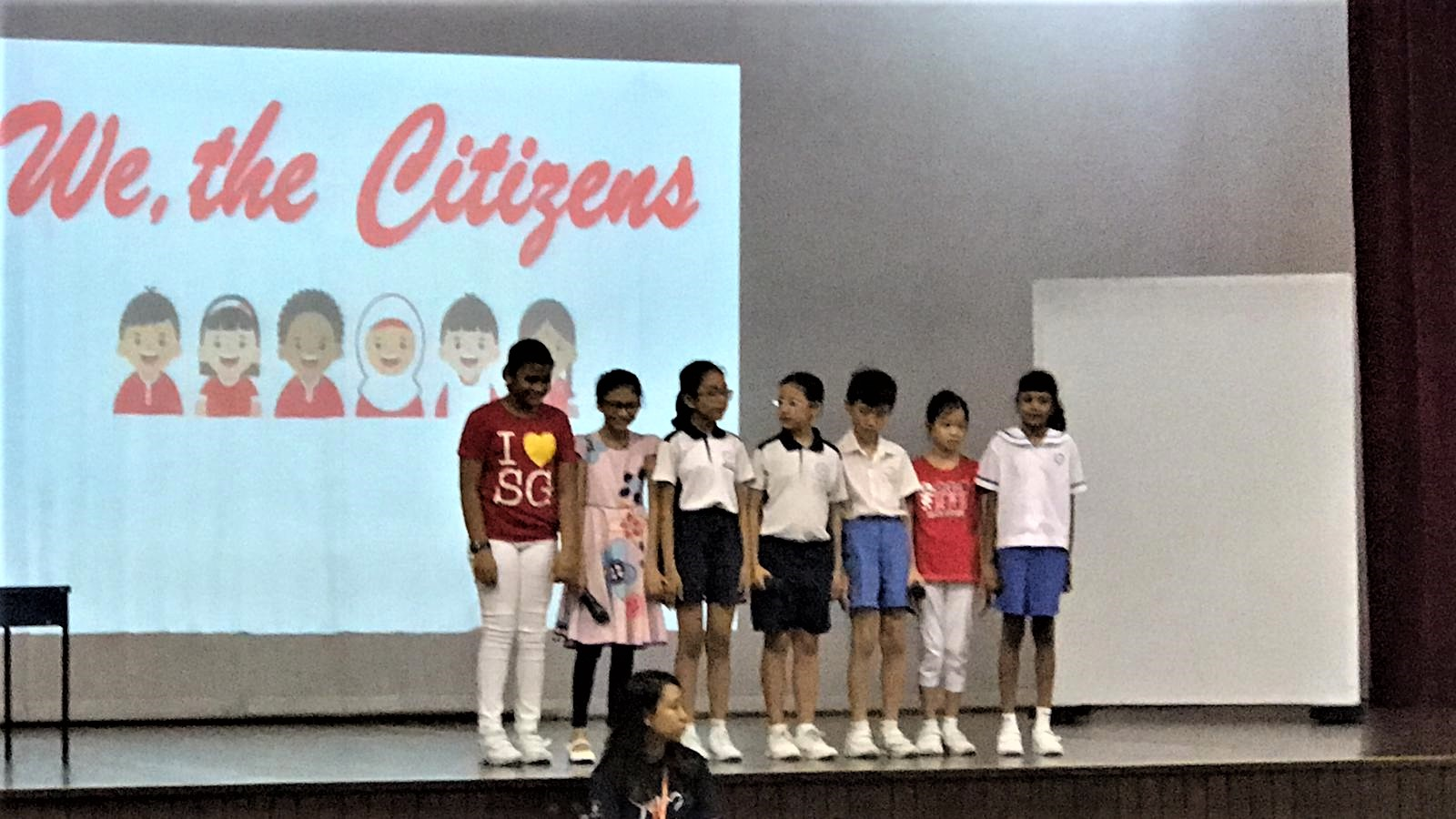 Seven children stand on a stage in front of a projection screen reading We, the Citizens.